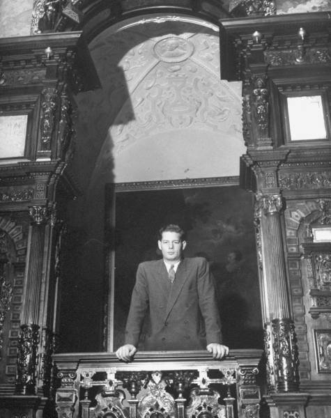 Rumanian King Michael leaning over balcony looking into large hall in Sinaia palace.